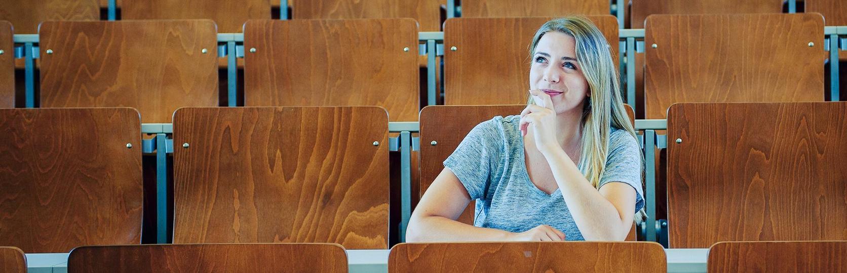 Female student sitting alone in a lecture hall with wooden chairs.